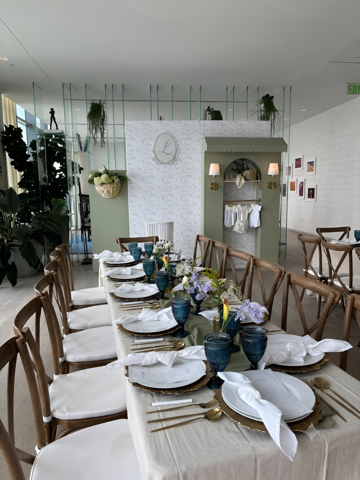 A long dining table set for guests in a bright, modern event space. The table is covered with a cream tablecloth and decorated with white plates, gold cutlery, blue glass vases, and floral centerpieces.