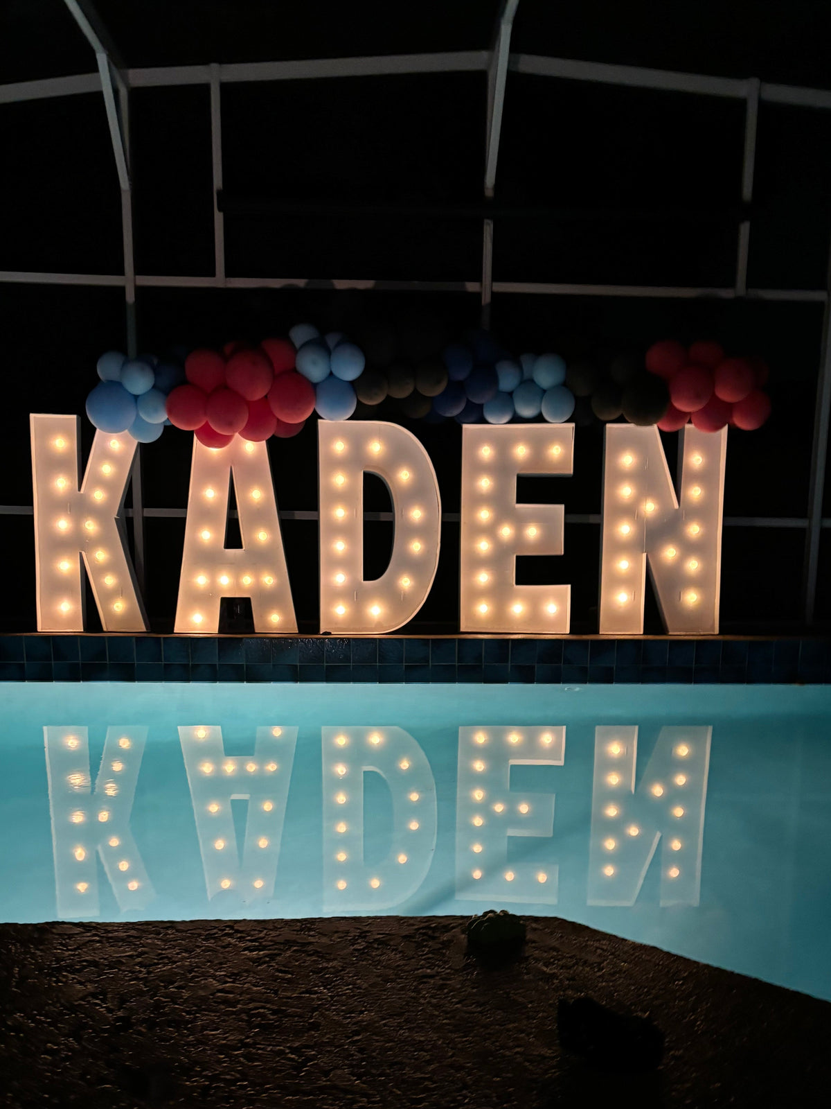 Large illuminated marquee letters spelling KADEN displayed on a pool deck at night, with balloons above and the letters reflected in the pool water.